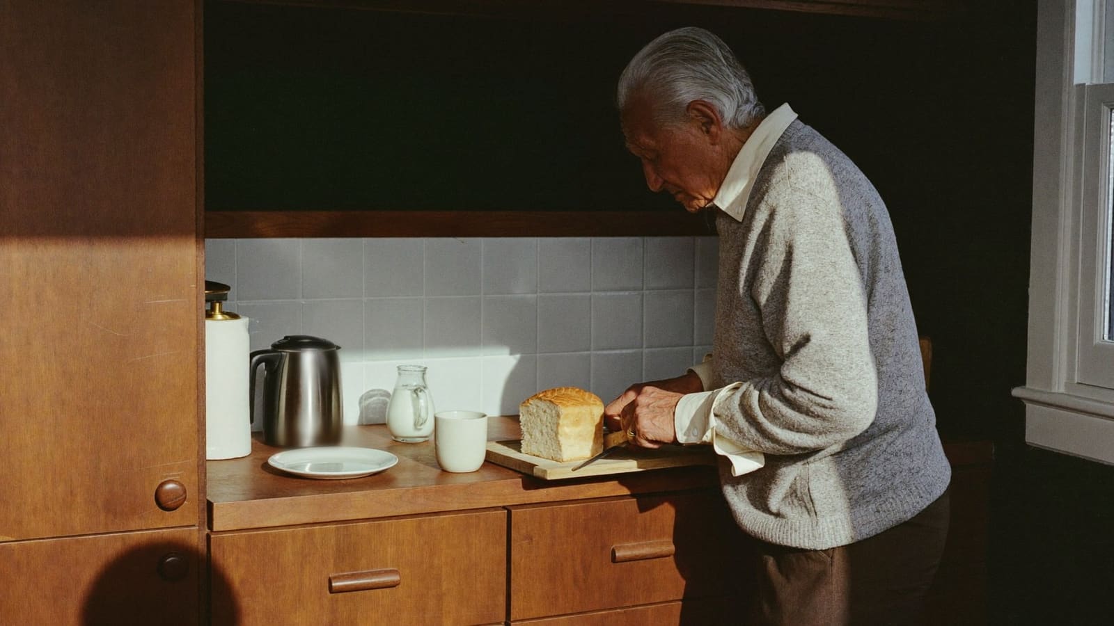An older man at his kitchen bench cutting bread for his own breakfast in the early morning light.