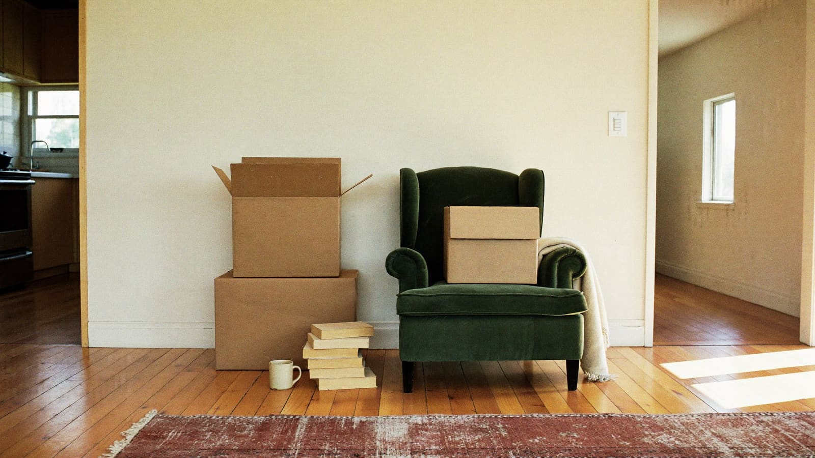 A part-packed lounge room, with an open brown cardboard carton stacked on a closed one against a cream wall, a third carton and a folded cream blanket on a worn dark green armchair, and a stack of books and a mug on the timber floorboards.