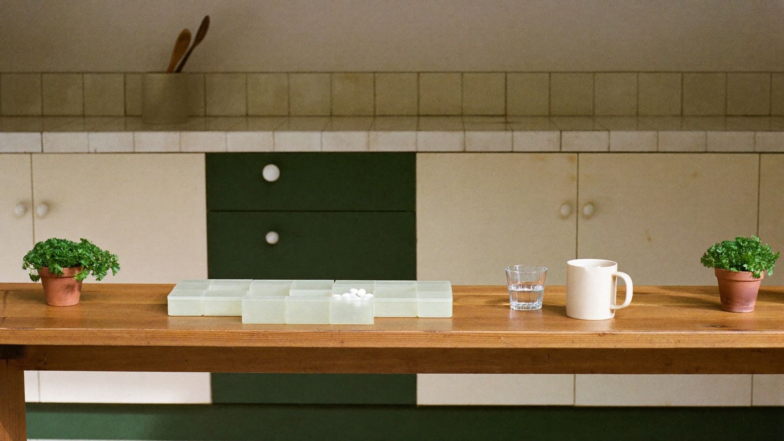 A row of small translucent lidded pill compartments lying on a honey-toned timber kitchen bench, one lid open with white tablets inside, beside a glass of water, a plain cream mug and two small terracotta pots of parsley.