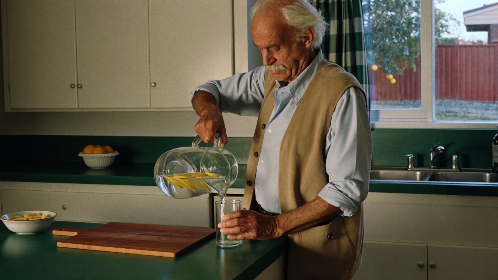 An older man in a tan vest pours water with lemon slices from a glass jug into a tumbler at his kitchen bench.