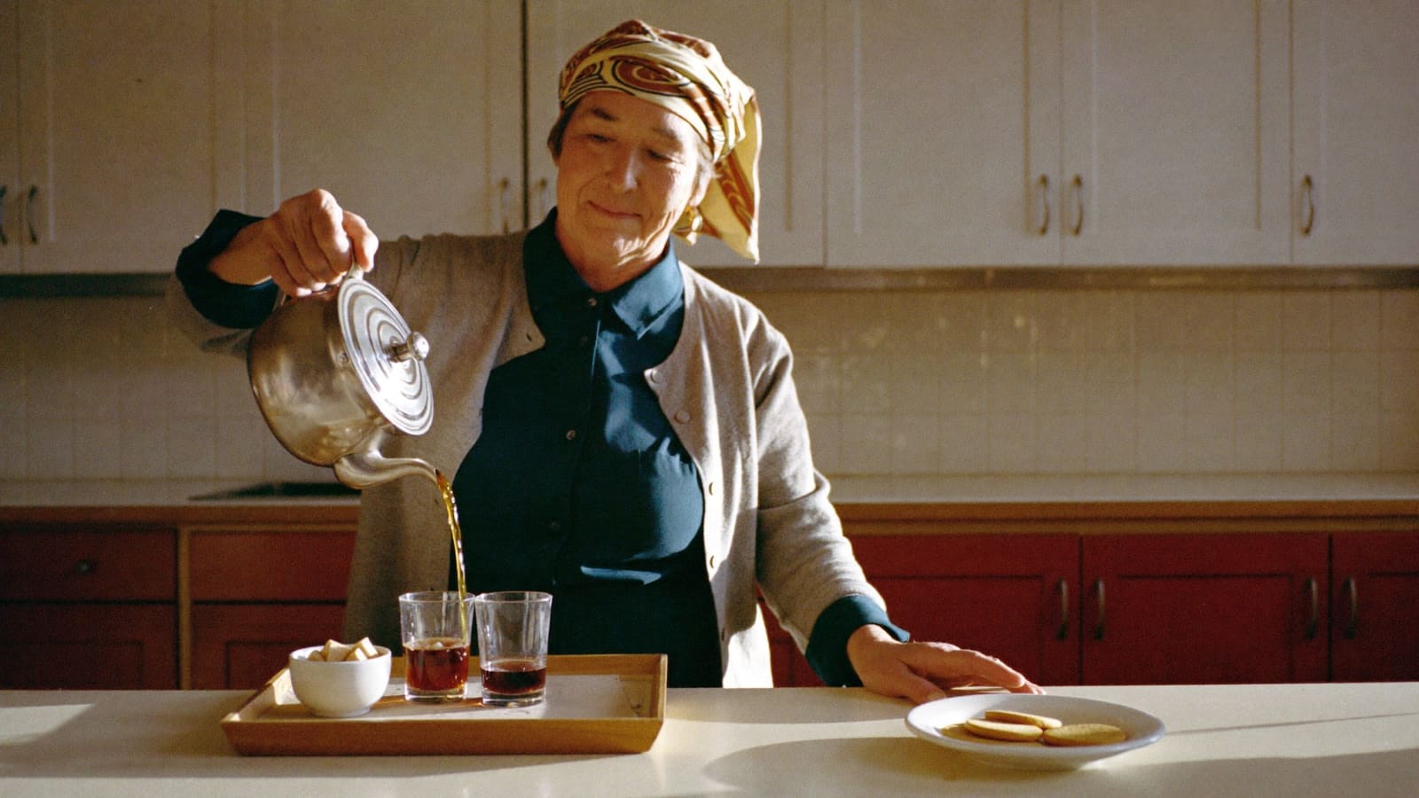 A woman in her seventies in a teal blouse, grey cardigan and patterned headscarf pours tea from a silver pot into two short glasses on a wooden tray at her kitchen bench, a plate of plain biscuits beside her.