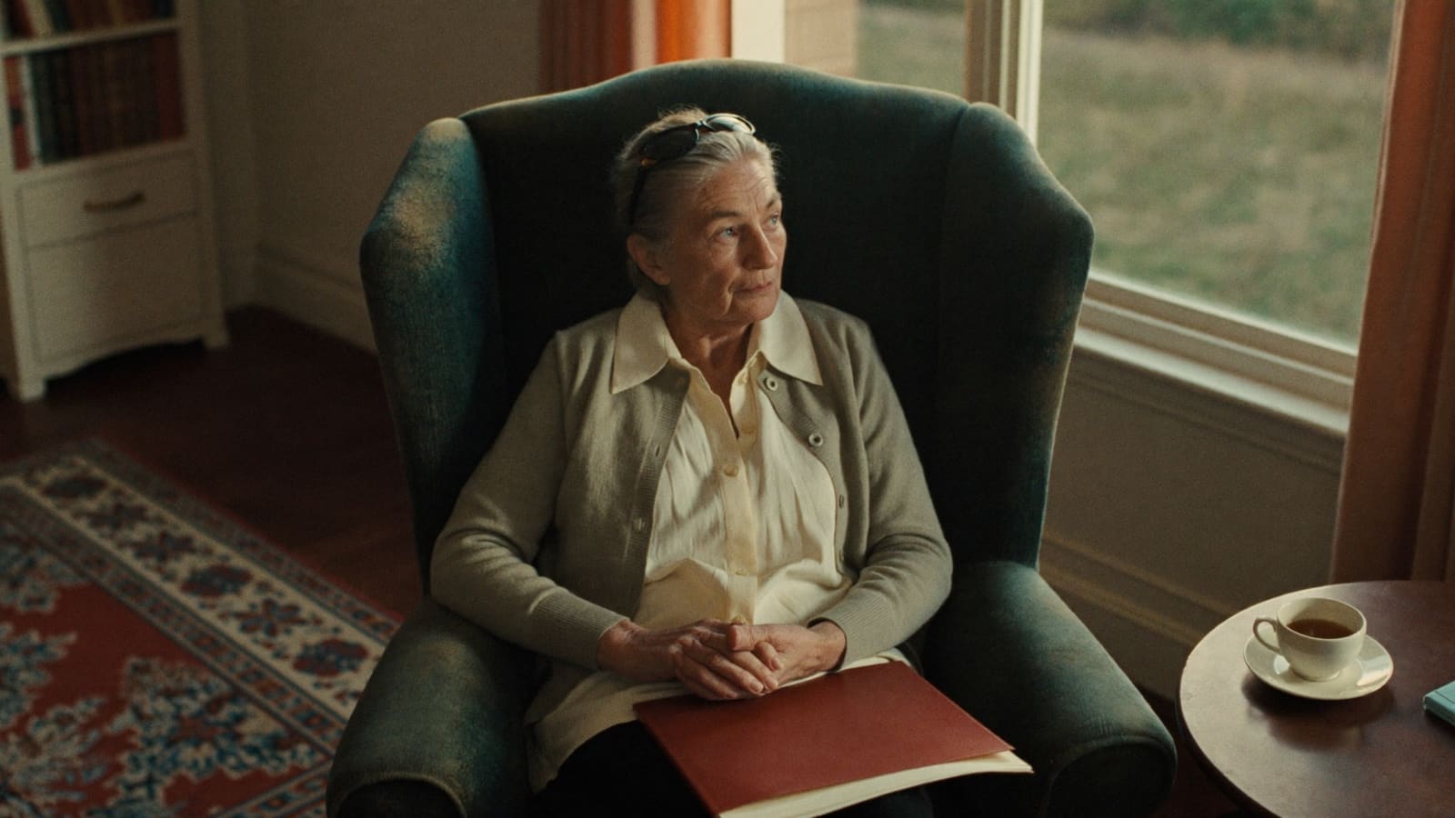An older woman in her armchair with a folder on her lap and her reading glasses pushed up, thinking something over.