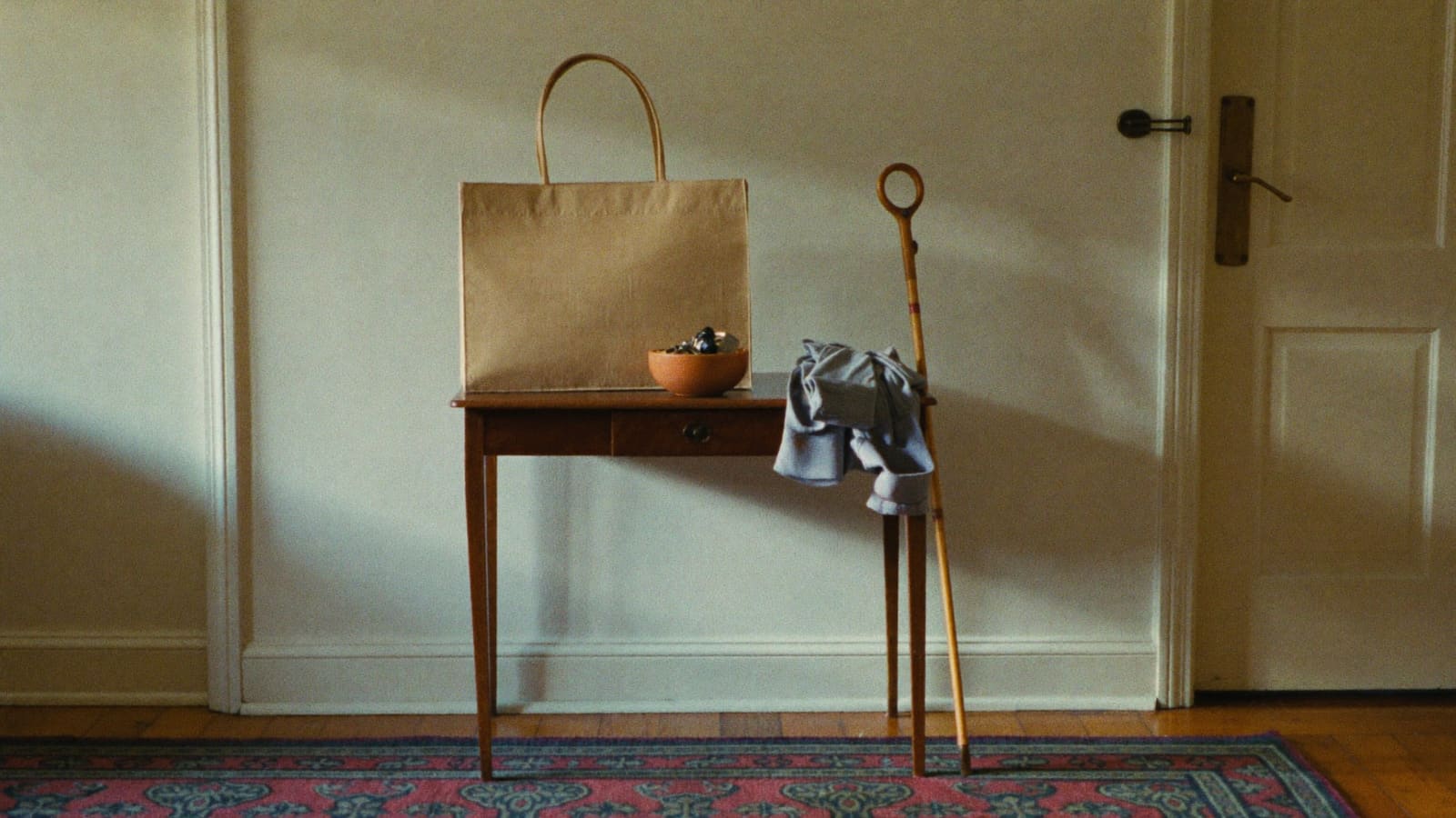 A hall table by a front door holding a plain canvas bag, keys in a terracotta bowl, a folded cardigan and a walking stick.