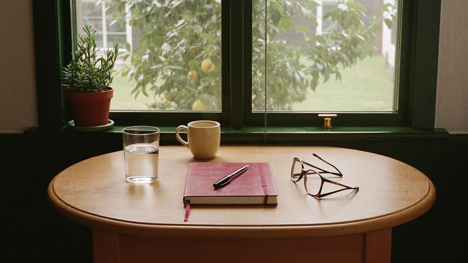 A closed deep red notebook with a pen on top, a glass of water, a cream mug and a pair of reading glasses on a round timber table by a window, with a pot of rosemary on the sill and a lemon tree outside.