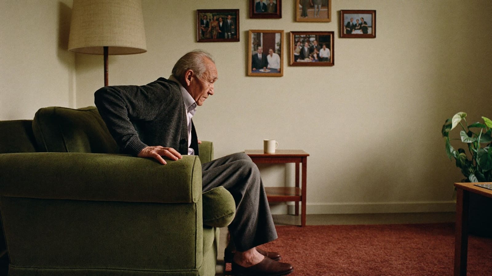 An older man rising steadily from an armchair in his own lounge room, both hands on the chair arms.