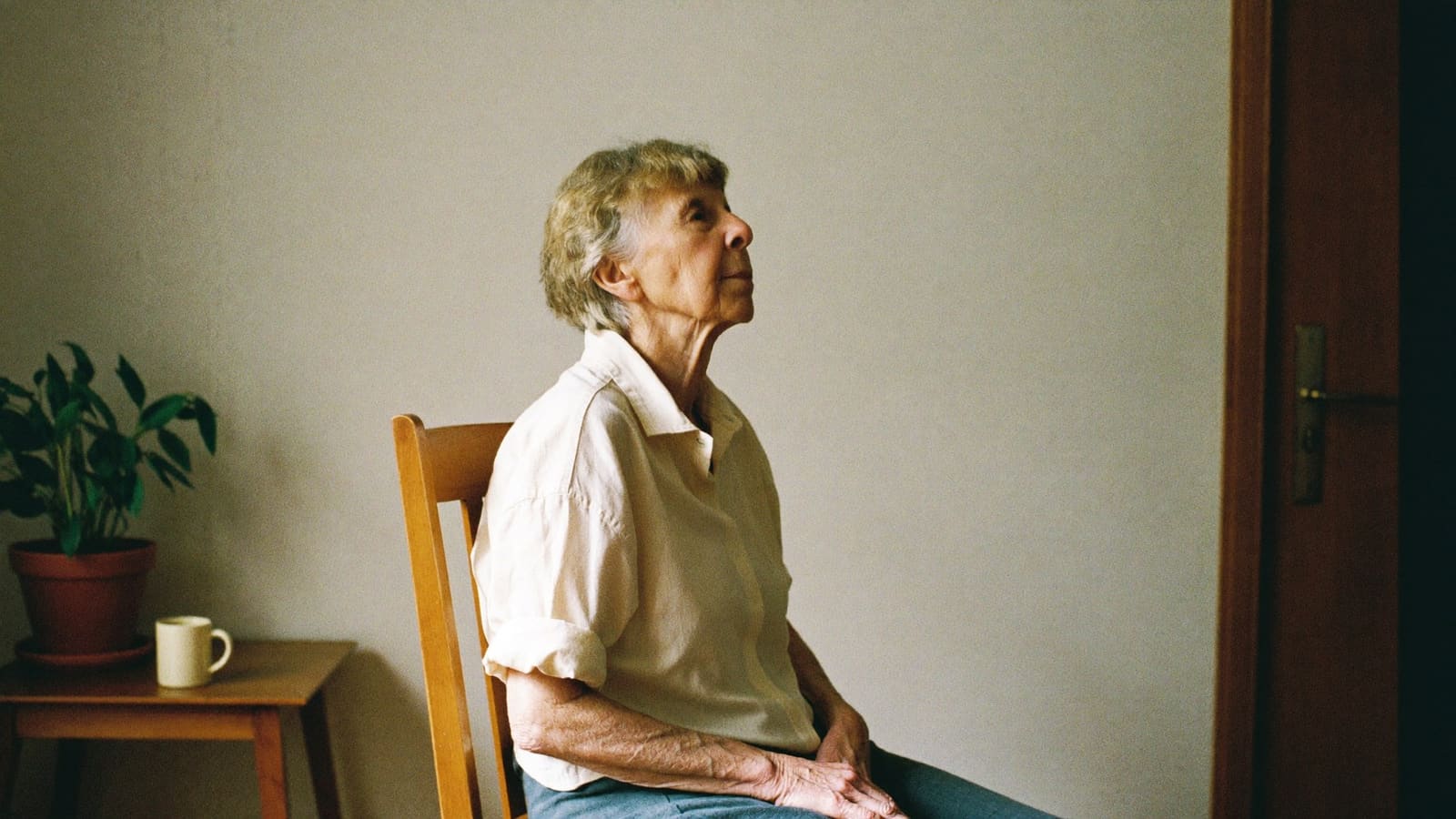 An older woman sitting upright and at ease on a dining chair in her living room.