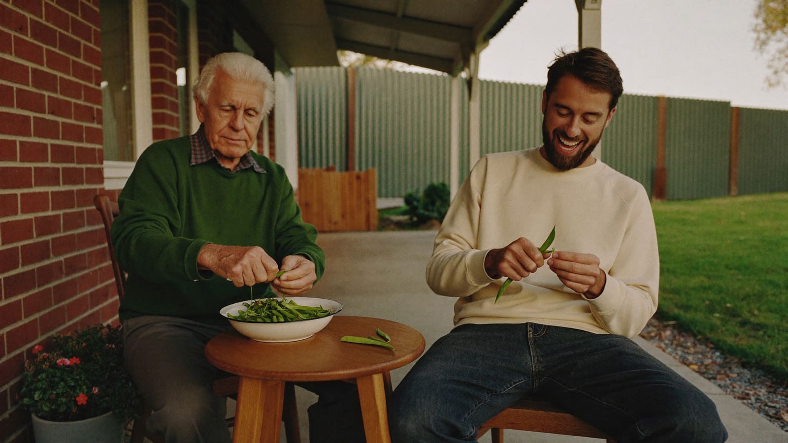 An older man and his adult grandson sit on a brick home's back veranda shelling peas into a white bowl, the grandson laughing.