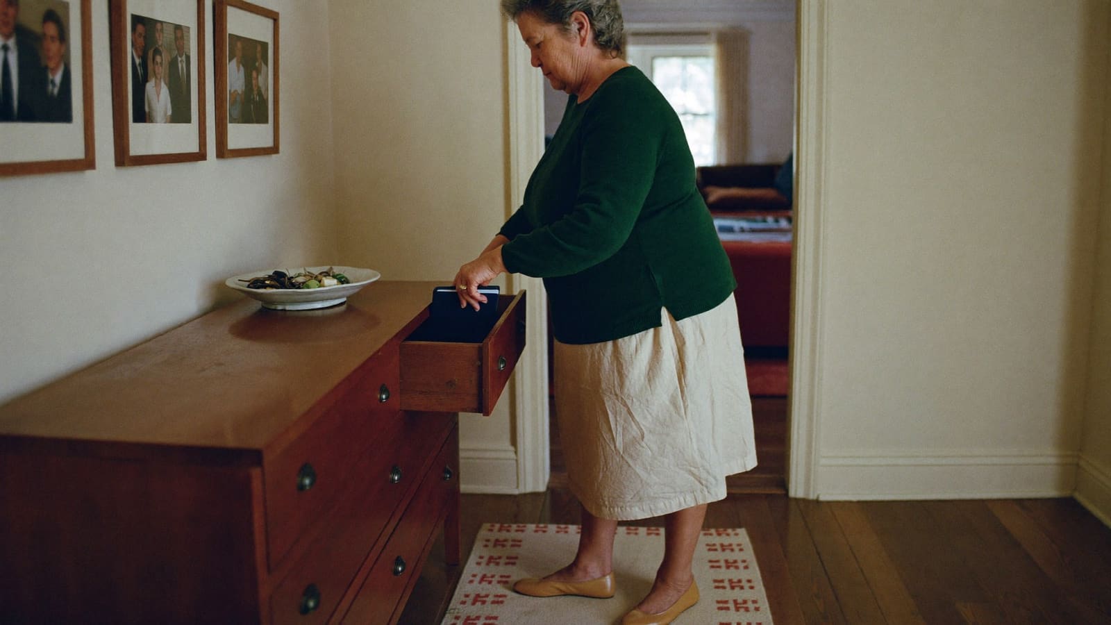 An older woman placing a plain document wallet into the drawer of a hallway sideboard.