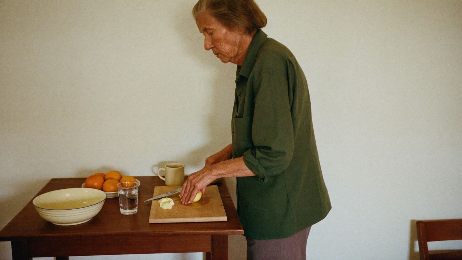 An older woman standing at her kitchen table slicing an apple, making herself something to eat.