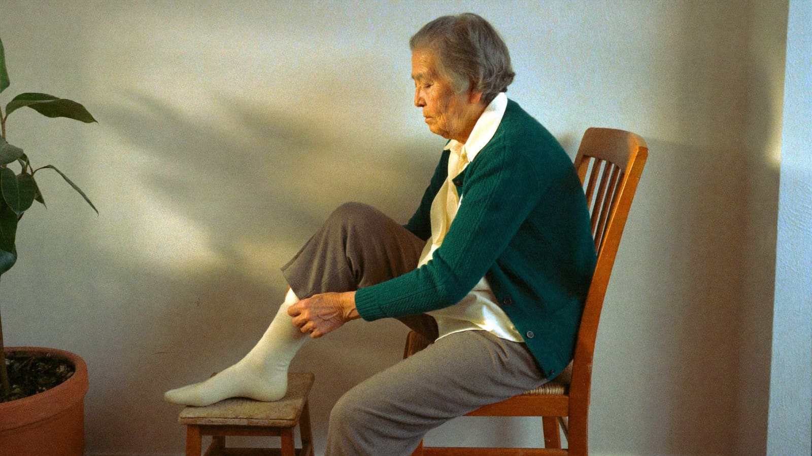 An older woman sitting on a kitchen chair with one foot resting on a low timber footstool, straightening her sock.