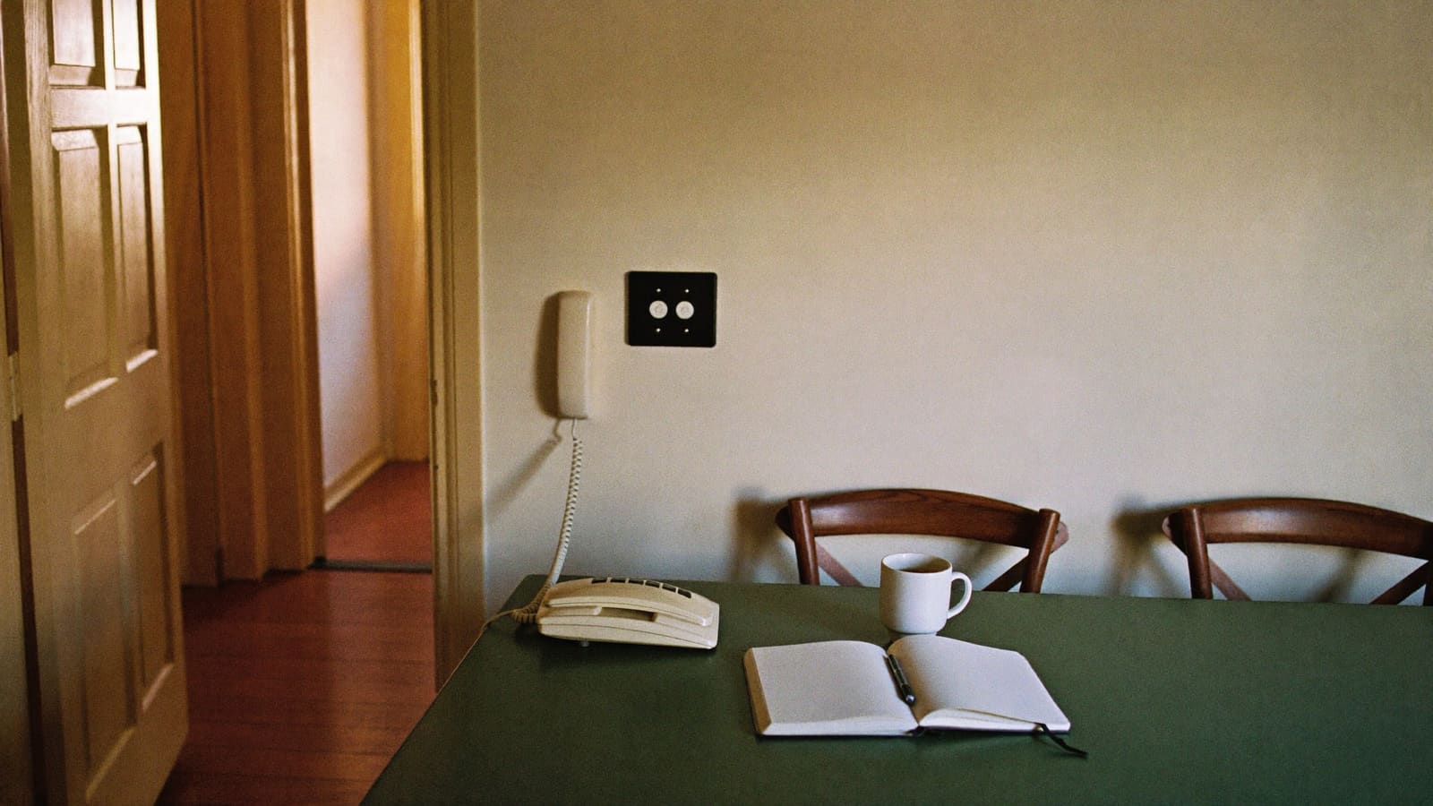 A telephone handset resting on a kitchen table beside an open notebook, a pen and a mug of tea, with two empty timber chairs.