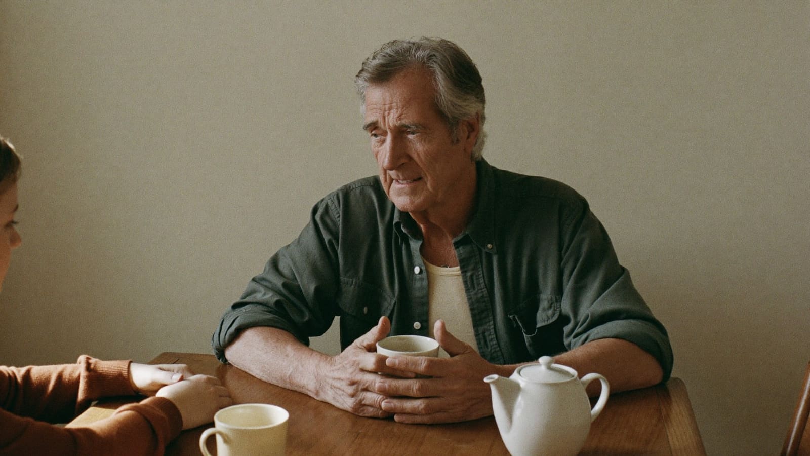 An older man in an olive shirt sits close to a timber kitchen table with both forearms on it and his hands around a cream mug, talking to a woman in a rust cardigan whose shoulder and hands are just visible at the edge of the frame.