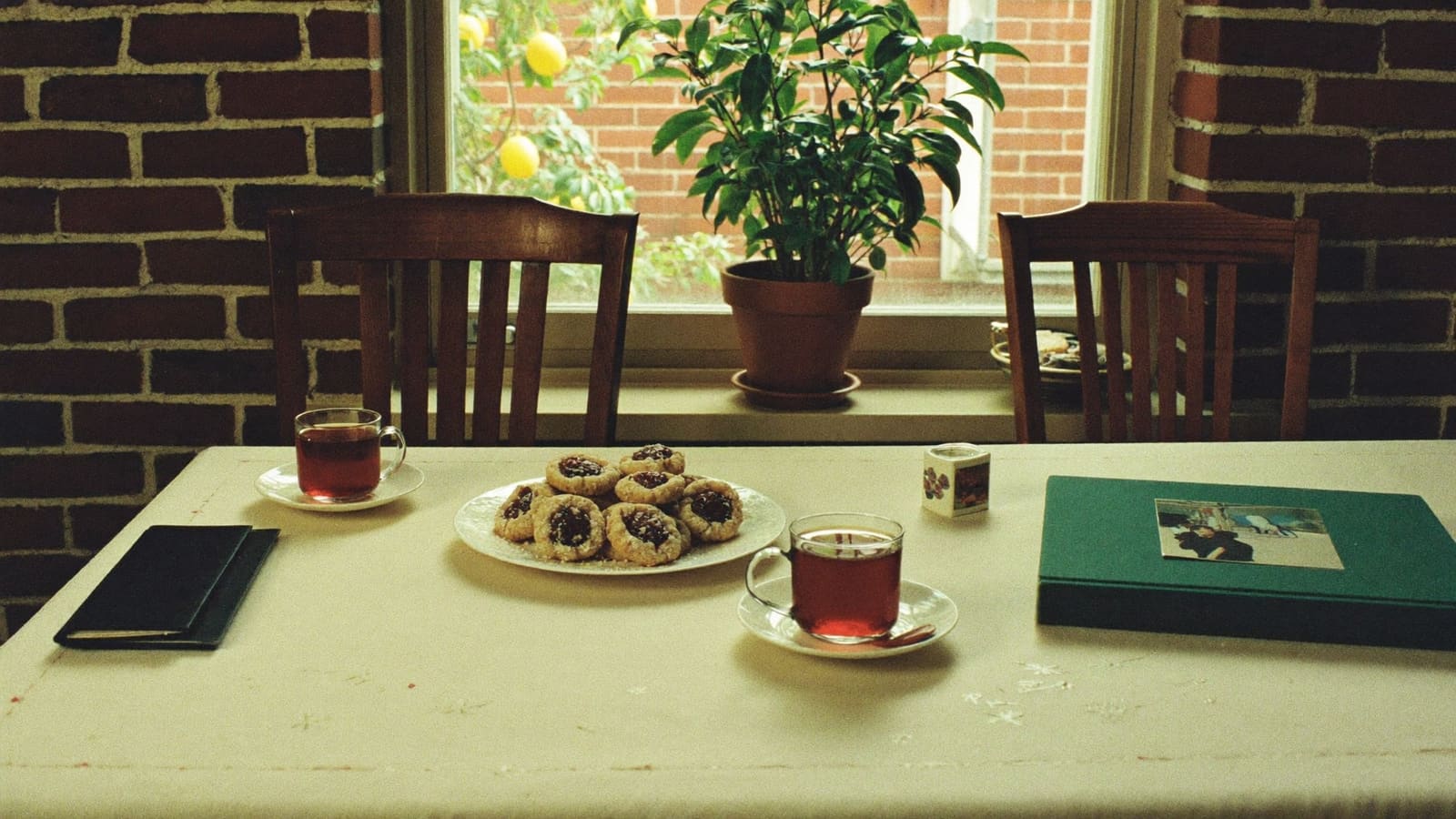 A kitchen table in a brick home set with two glasses of black tea on saucers, a plate of homemade biscuits and a closed green photo album with a small family snapshot on the cover, two chairs pulled out.
