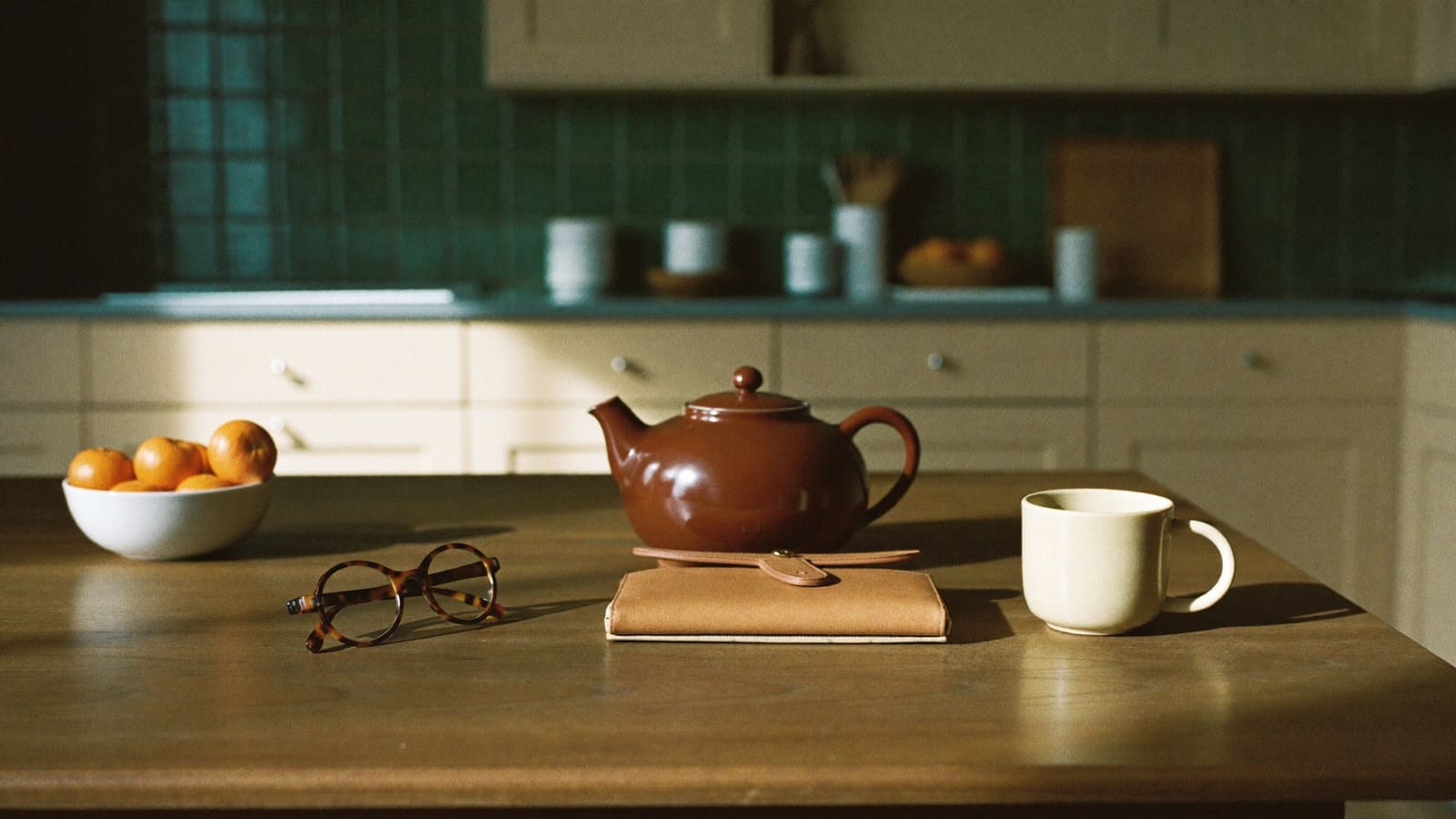 A brown teapot, a closed tan leather wallet, a pair of tortoiseshell reading glasses, a cream mug and a bowl of mandarins on a timber kitchen table with a green tiled kitchen behind.