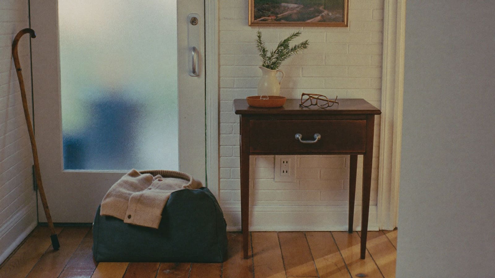 A packed travel bag and a folded cardigan by the front door of a suburban home, with keys in a bowl on the hall table.