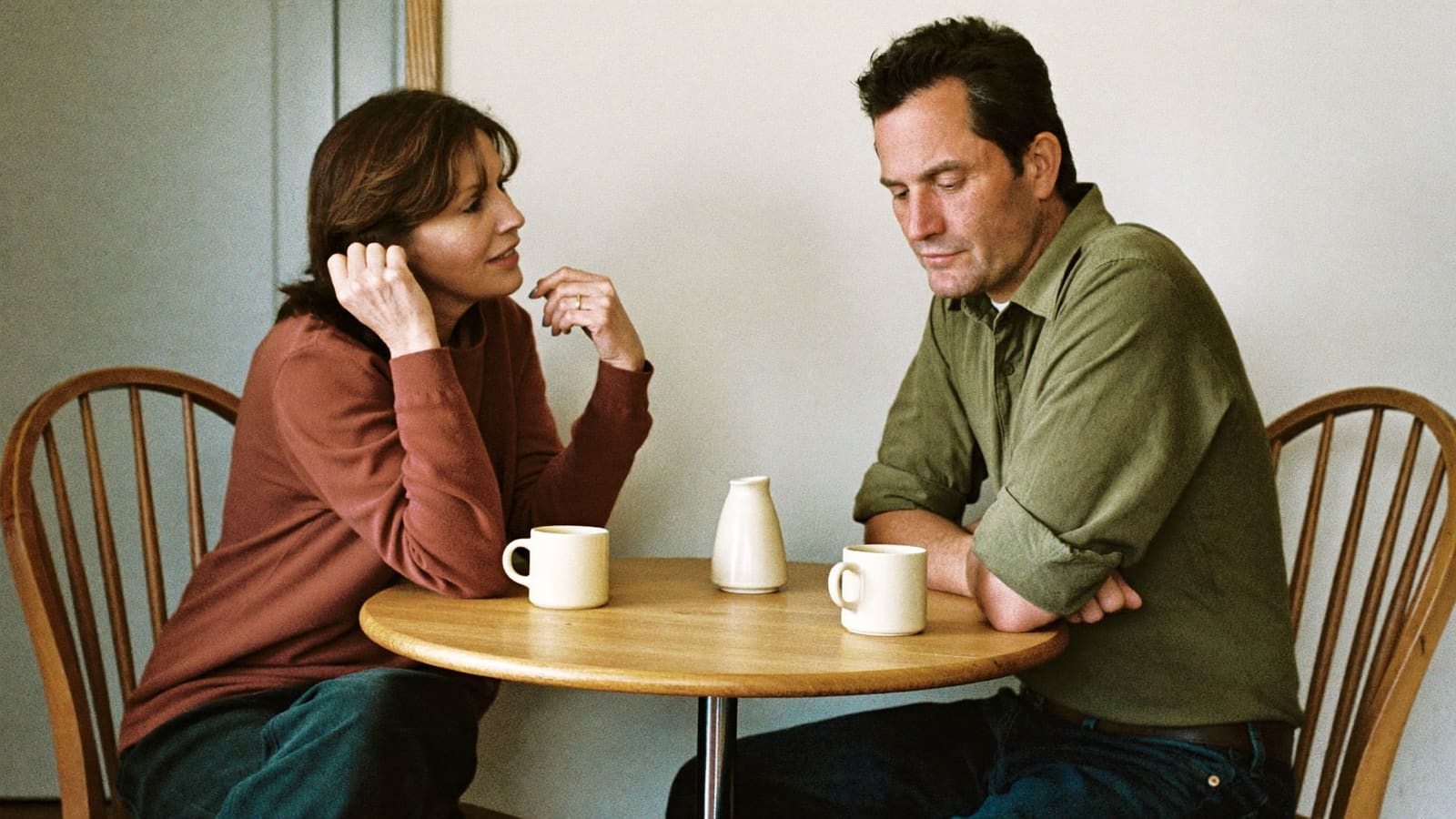 Two adult siblings sitting close at a round kitchen table over mugs of tea, talking.