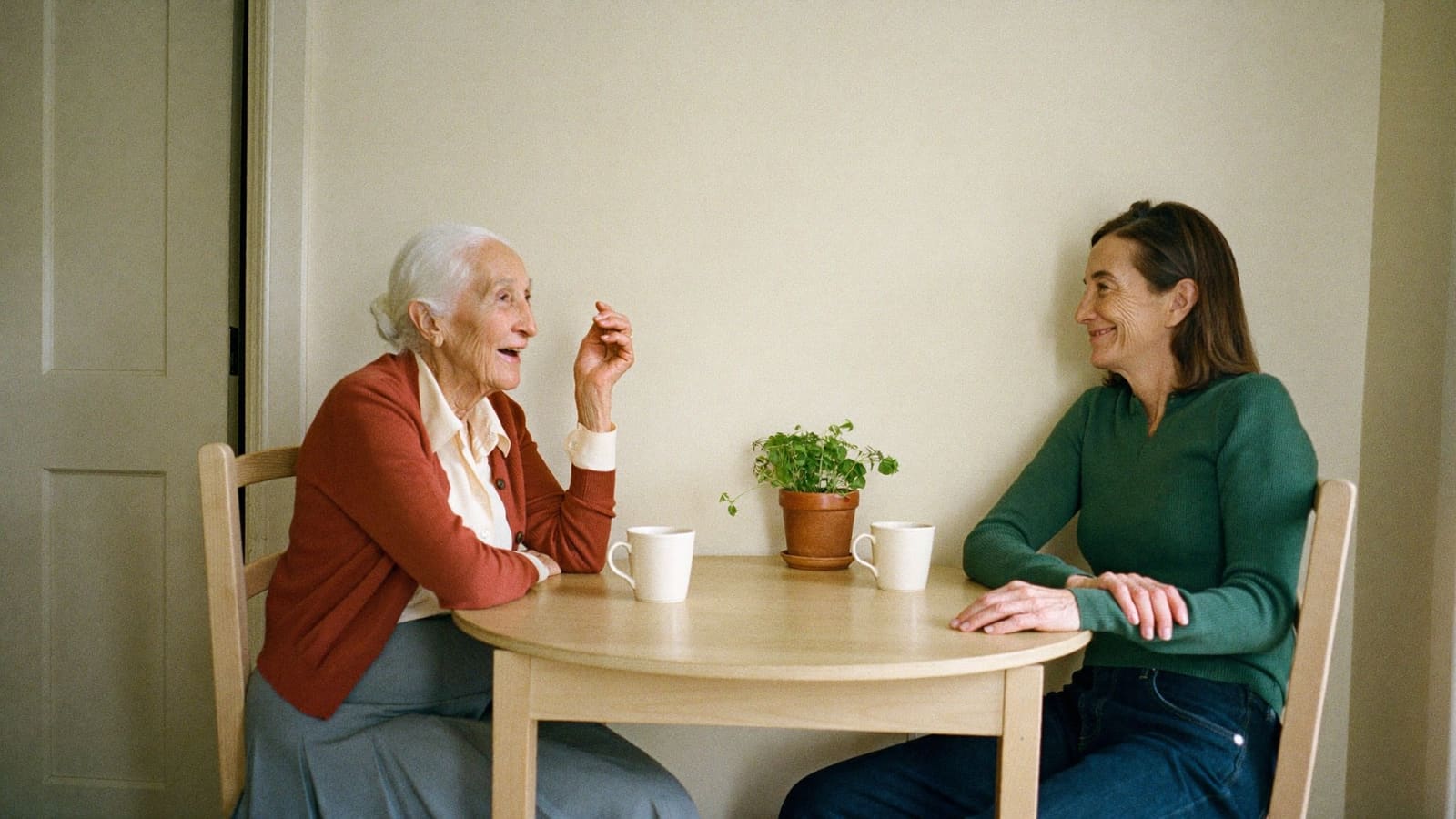 Two women sitting close at a small kitchen table, leaning in and talking easily with each other.