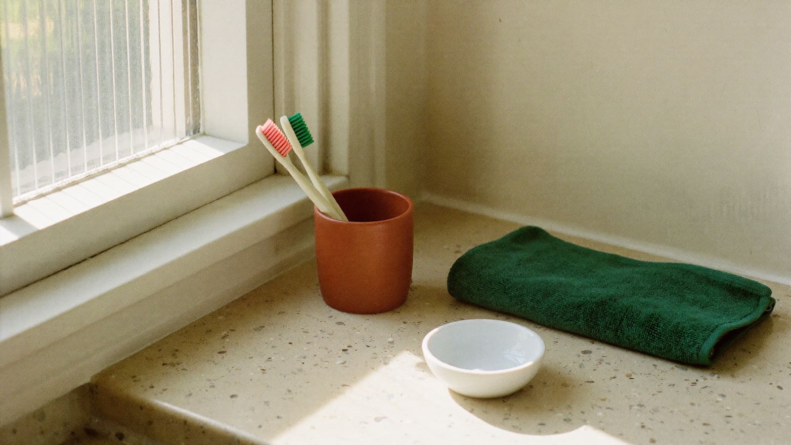 A terracotta ceramic cup holding two soft toothbrushes on a pale terrazzo bathroom bench, beside a folded deep green hand towel and a small white dish, in soft light from a frosted window.