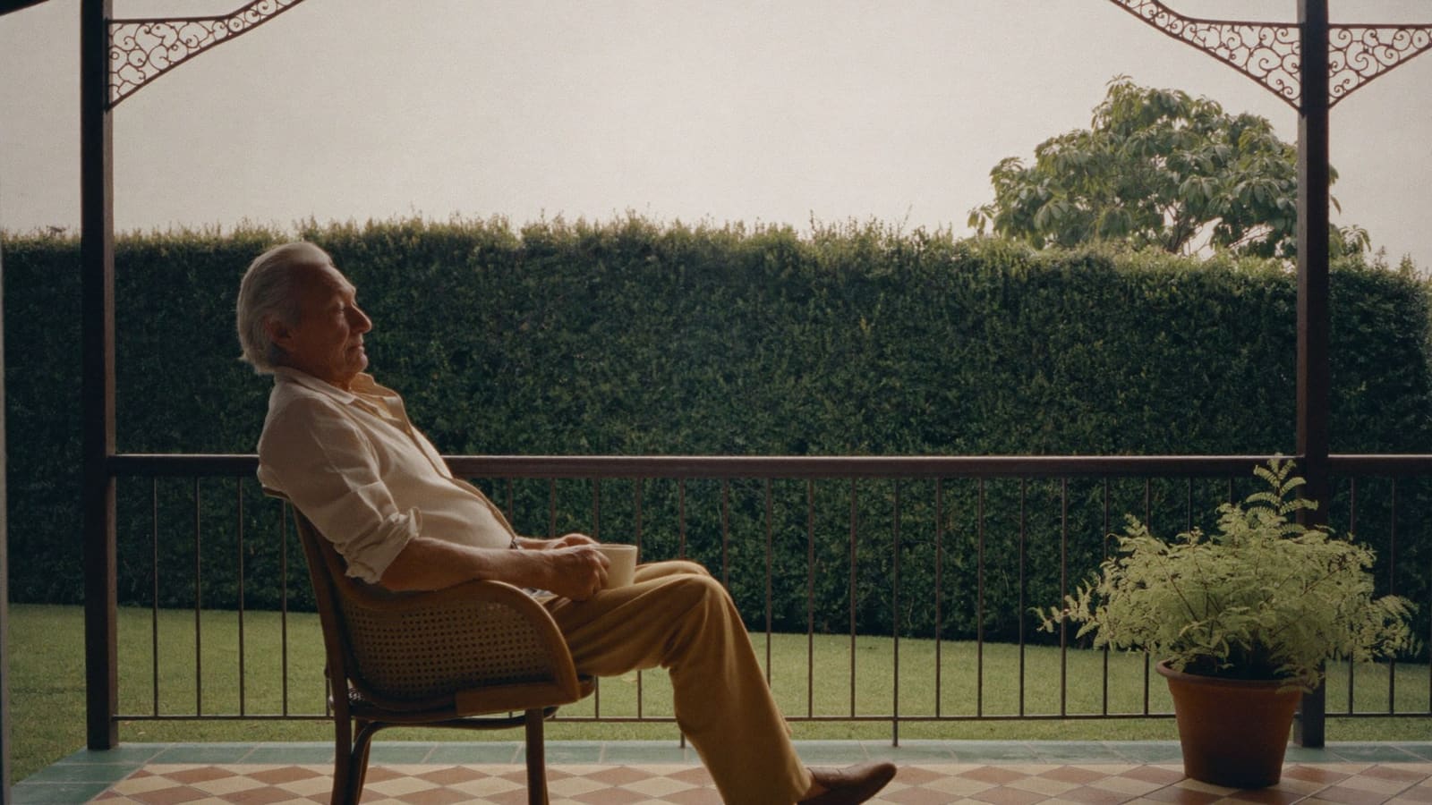 An older man sitting in a cane chair on his own front veranda with a mug of tea in both hands.