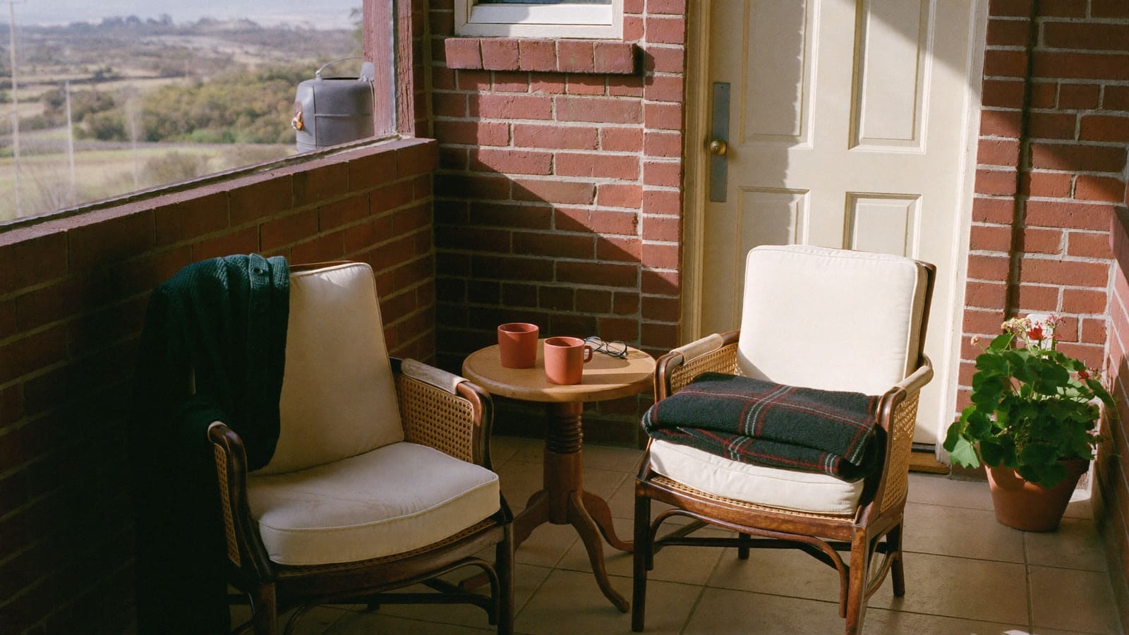Two cane armchairs with cream cushions on a sunny brick back veranda, a green cardigan over one and a tartan rug on the other, two terracotta mugs and a pair of reading glasses on the small table between them.