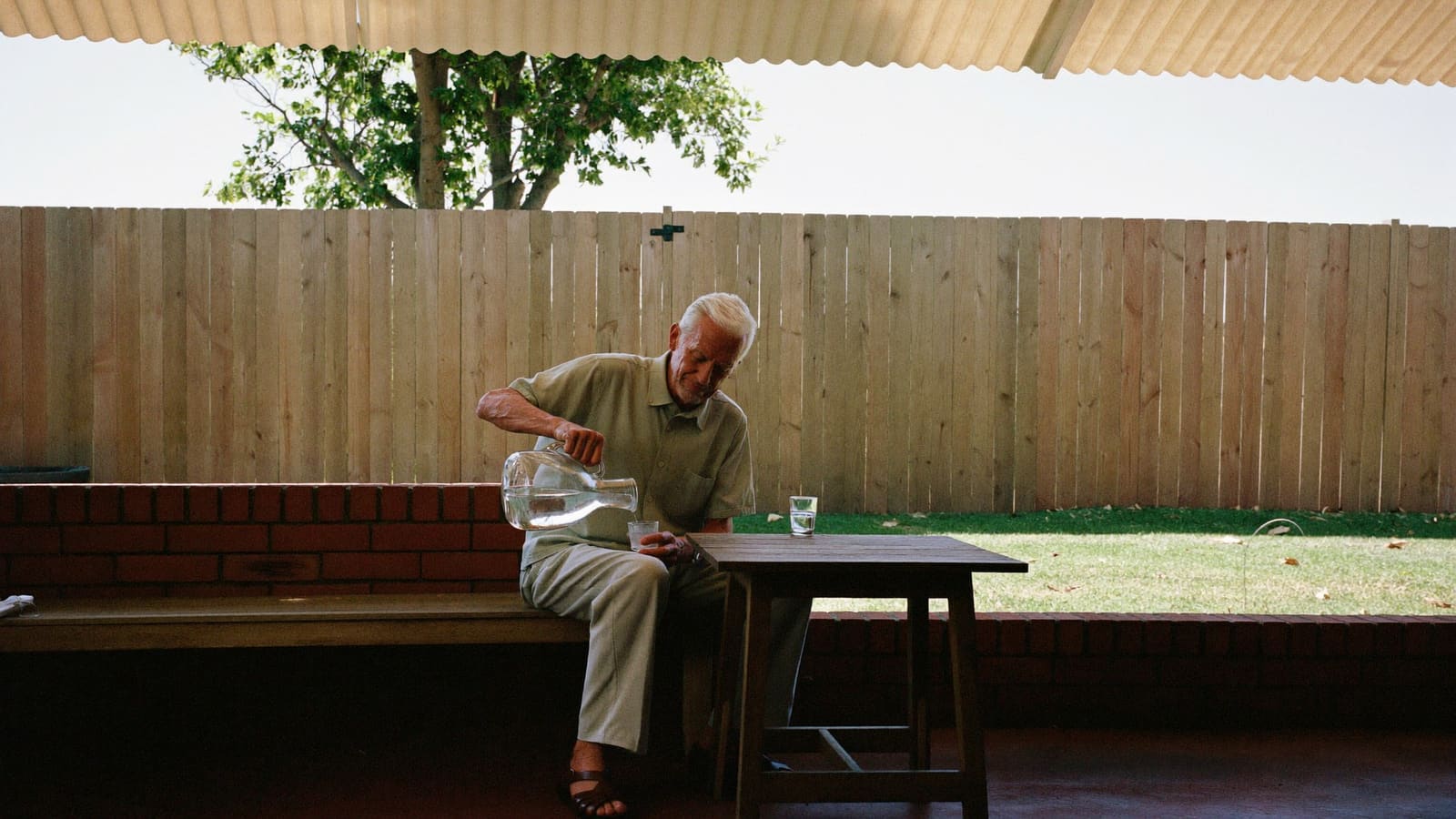 An older man sitting in deep shade on his back veranda pouring a glass of water on a hot day.