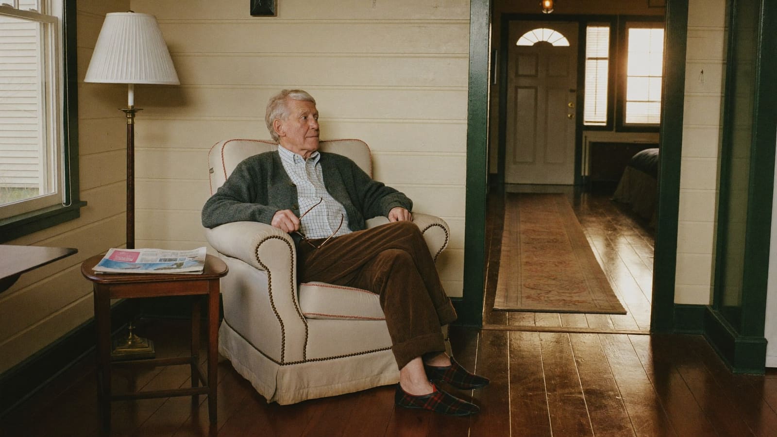 An older man in an armchair in his front room, glasses in hand, looking down the hallway toward the front door.